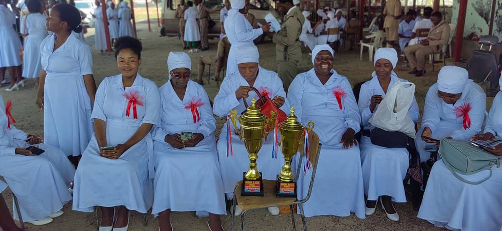 Choir members in white seated beside two gold trophies on a chair