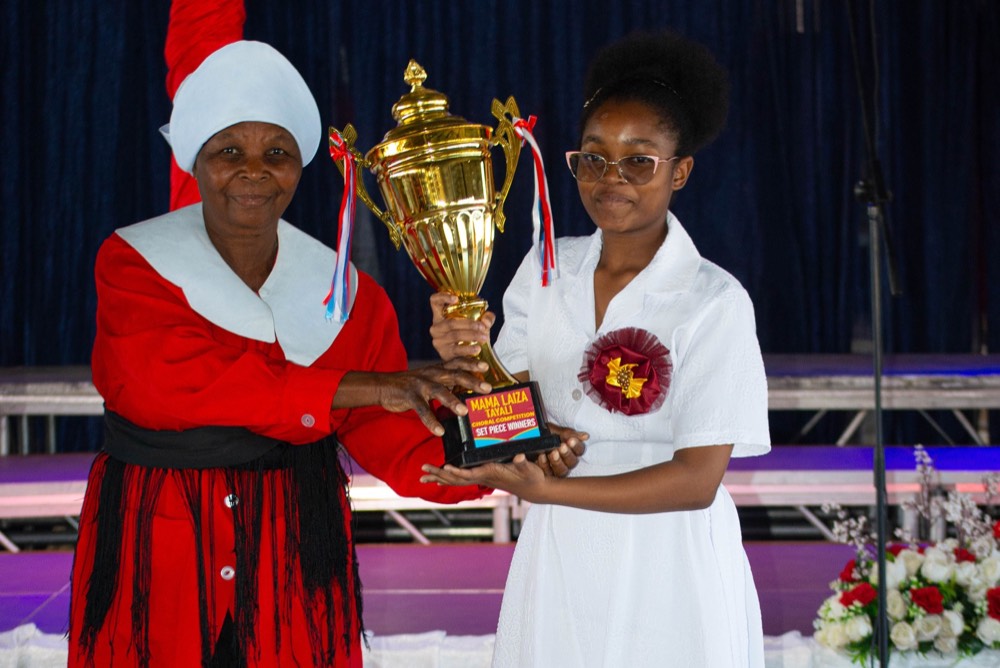 Two women on stage holding a gold trophy labelled Mama Laiza Tayali Choral Competition, Set Piece Winners